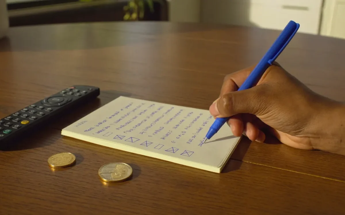 A hand ticks the last box on a five-point paper checklist beside a TV remote and coins, the safe way to buy IPTV in Canada